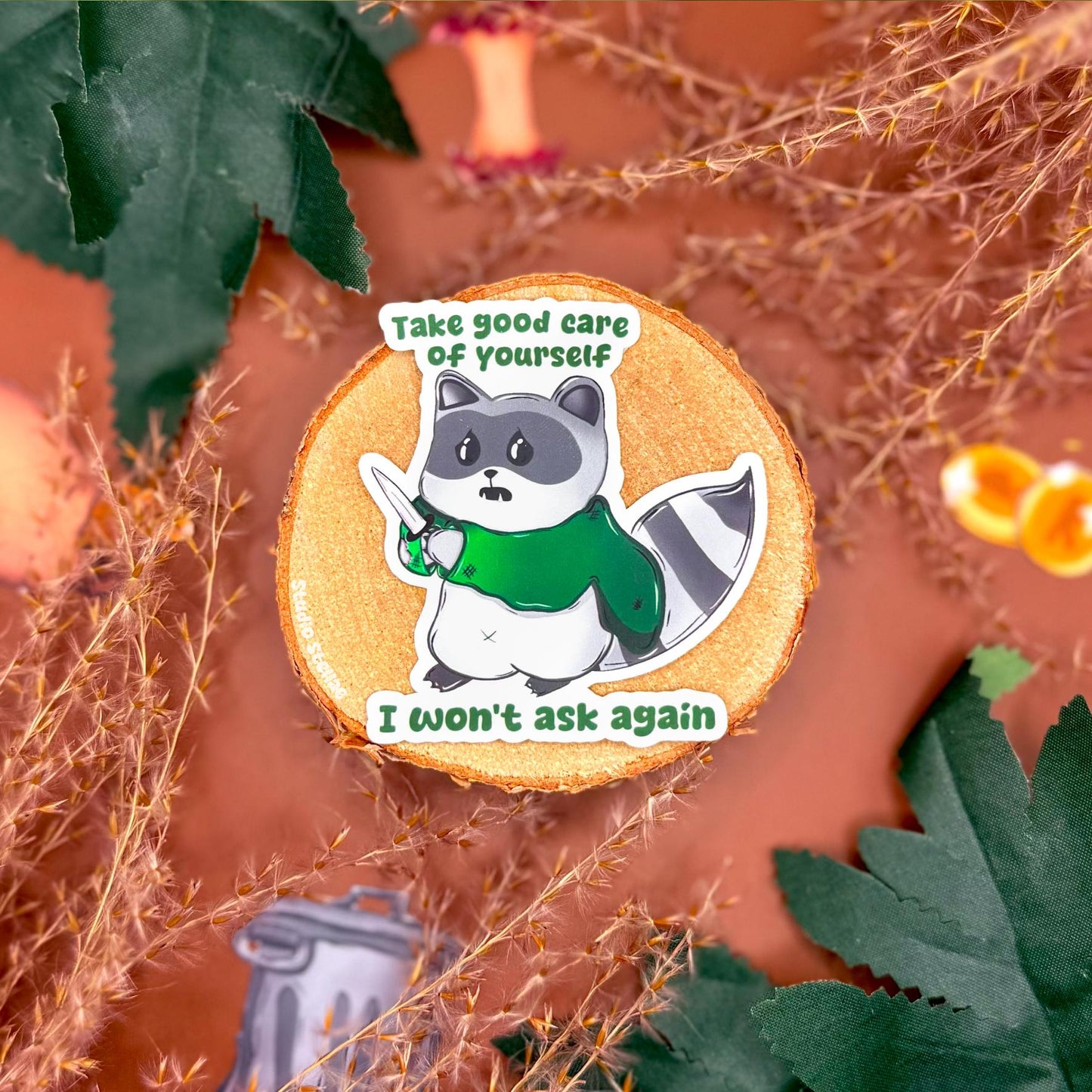 Self care raccoon sticker displayed on a warm autumn styled background with leaves and cozy props, featuring a cute trash panda holding a knife with the quote “Take good care of yourself, I won’t ask again.” This funny mental health raccoon sticker highlights its matte finish and playful design, showing a sarcastic self care reminder decal perfect for laptops, planners, journals, or water bottles and a relatable gift for anyone who loves cute but chaotic animal humor.