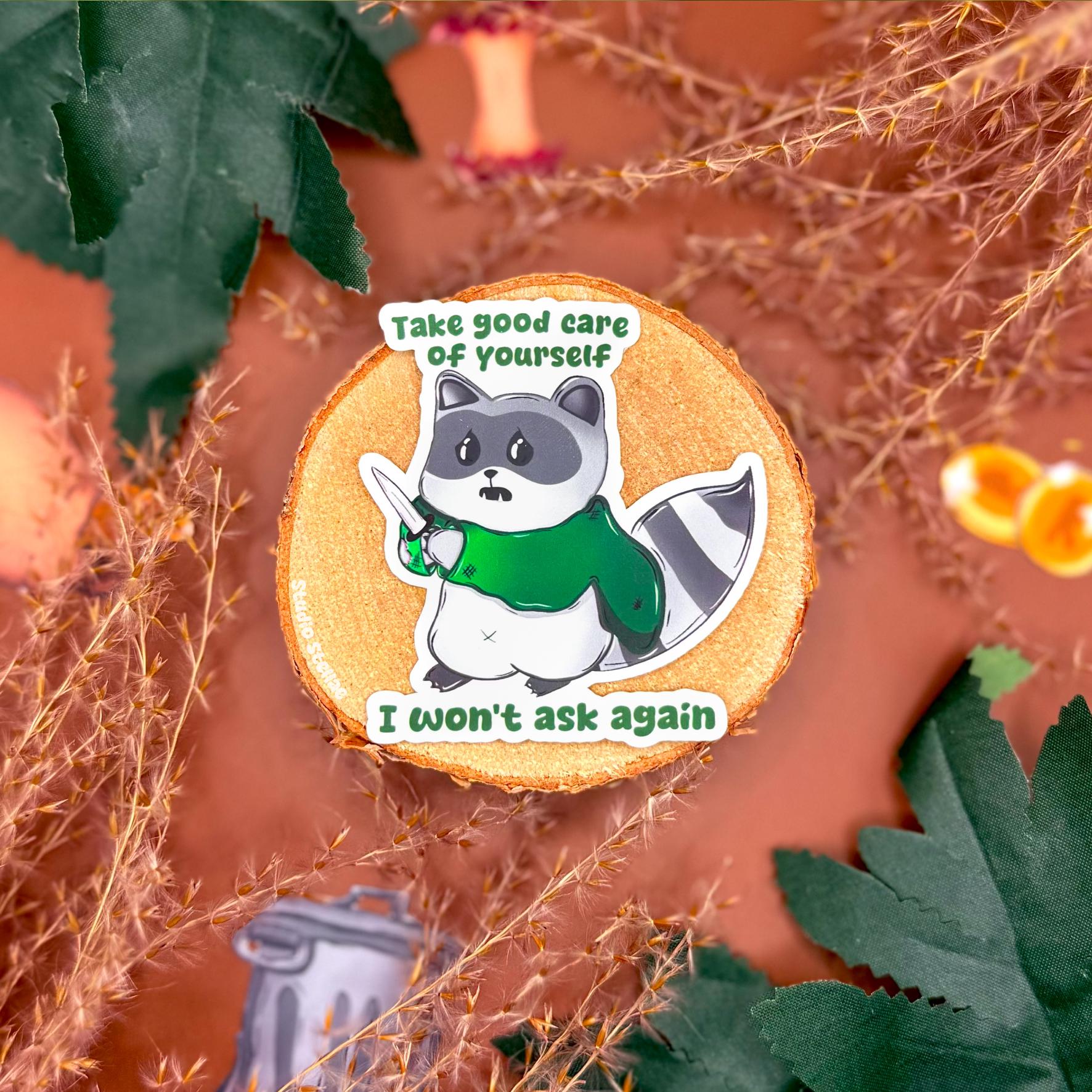 Self care raccoon sticker displayed on a warm autumn styled background with leaves and cozy props, featuring a cute trash panda holding a knife with the quote “Take good care of yourself, I won’t ask again.” This funny mental health raccoon sticker highlights its matte finish and playful design, showing a sarcastic self care reminder decal perfect for laptops, planners, journals, or water bottles and a relatable gift for anyone who loves cute but chaotic animal humor.