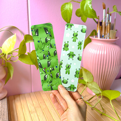 Hand holding the green alien bookmark set in a cozy pastel workspace with a pink wall, leafy green plants, and a pink vase filled with paintbrushes. Both bookmarks are visible, showing their bright green alien designs and smooth matte lamination. This lifestyle photo shows real-life scale in hand and how the bookmarks look in natural light, perfect for book lovers, journaling, school supplies, and gifting.