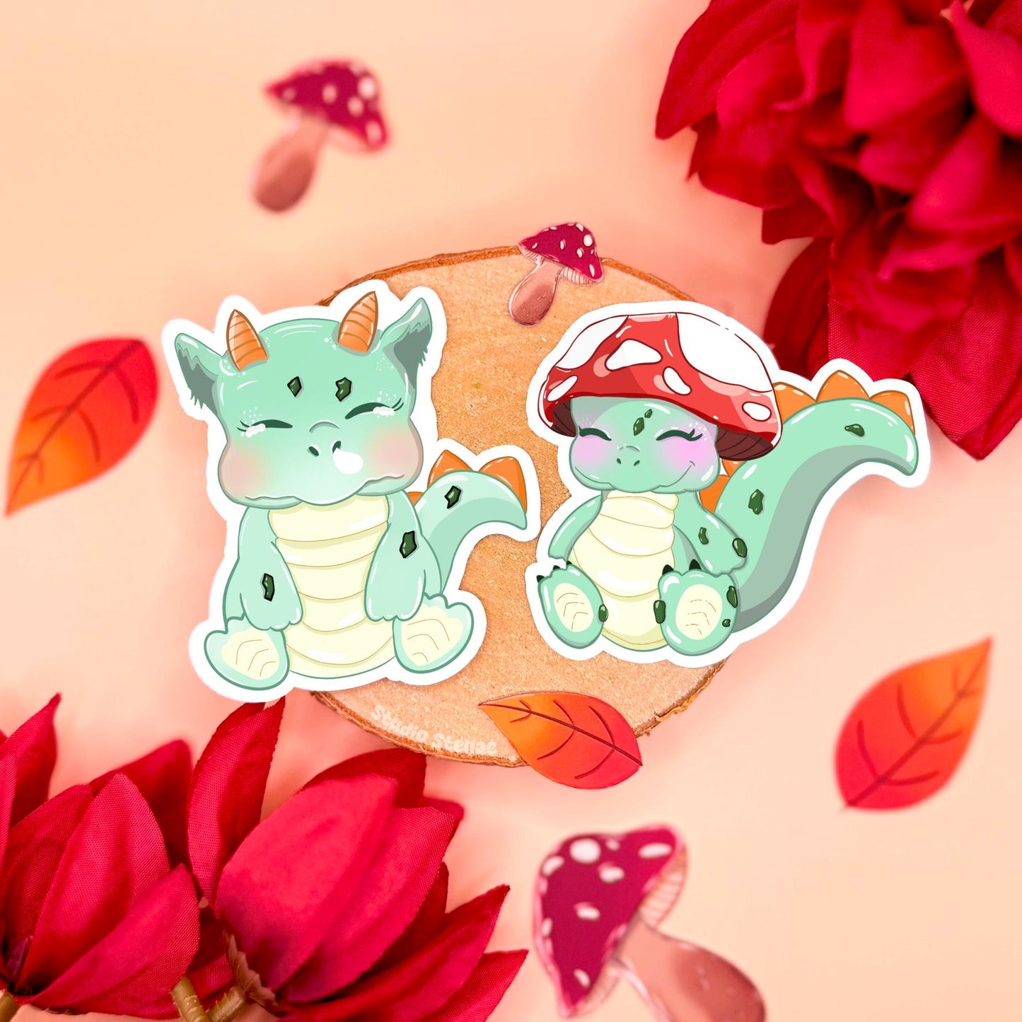 Cute baby dinosaur sticker set styled in a soft pastel flat lay with cork, red flowers and cozy cottagecore props, showing both chibi dinos side by side. One sleepy mint green dinosaur sits calmly while the smaller friend wears an adorable red mushroom hat. These kawaii cottagecore dino stickers are matte laminated, waterproof and perfect for decorating planners, journals, laptops and water bottles, making a sweet fantasy stationery gift for cute trinket lovers.