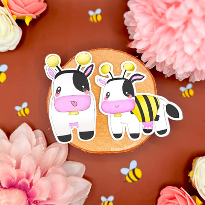 Bee cow sticker set styled on a warm brown background with pink flowers and tiny bee confetti, showing two kawaii chibi cows dressed as bumblebees. The cozy farm vibe and soft colors match MySims Kingdom nostalgia and cottagecore gamer aesthetics. This cute cow sticker set highlights crisp die cut edges and matte laminated finish, perfect for decorating laptops, planners, journals, phone cases, or water bottles as a cozy gamer gift.