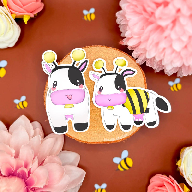 Bee cow sticker set styled on a warm brown background with pink flowers and tiny bee confetti, showing two kawaii chibi cows dressed as bumblebees. The cozy farm vibe and soft colors match MySims Kingdom nostalgia and cottagecore gamer aesthetics. This cute cow sticker set highlights crisp die cut edges and matte laminated finish, perfect for decorating laptops, planners, journals, phone cases, or water bottles as a cozy gamer gift.