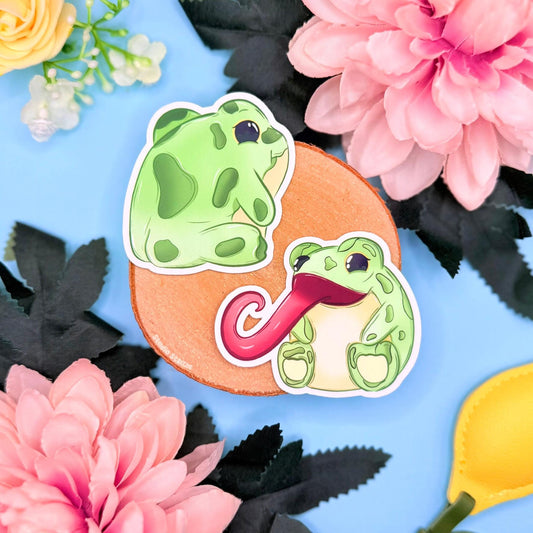 Cute frog sticker set styled in a soft pastel flat lay with cork board, flowers and greenery, showing both kawaii frog designs together. One silly green frog sticks out an extra long tongue while the other shows an adorable froggy bum with a cheeky smile. These funny handmade animal decals are matte laminated, waterproof and perfect for decorating laptops, planners, journals and water bottles, making a playful stationery gift for frog lovers and cute sticker collectors.