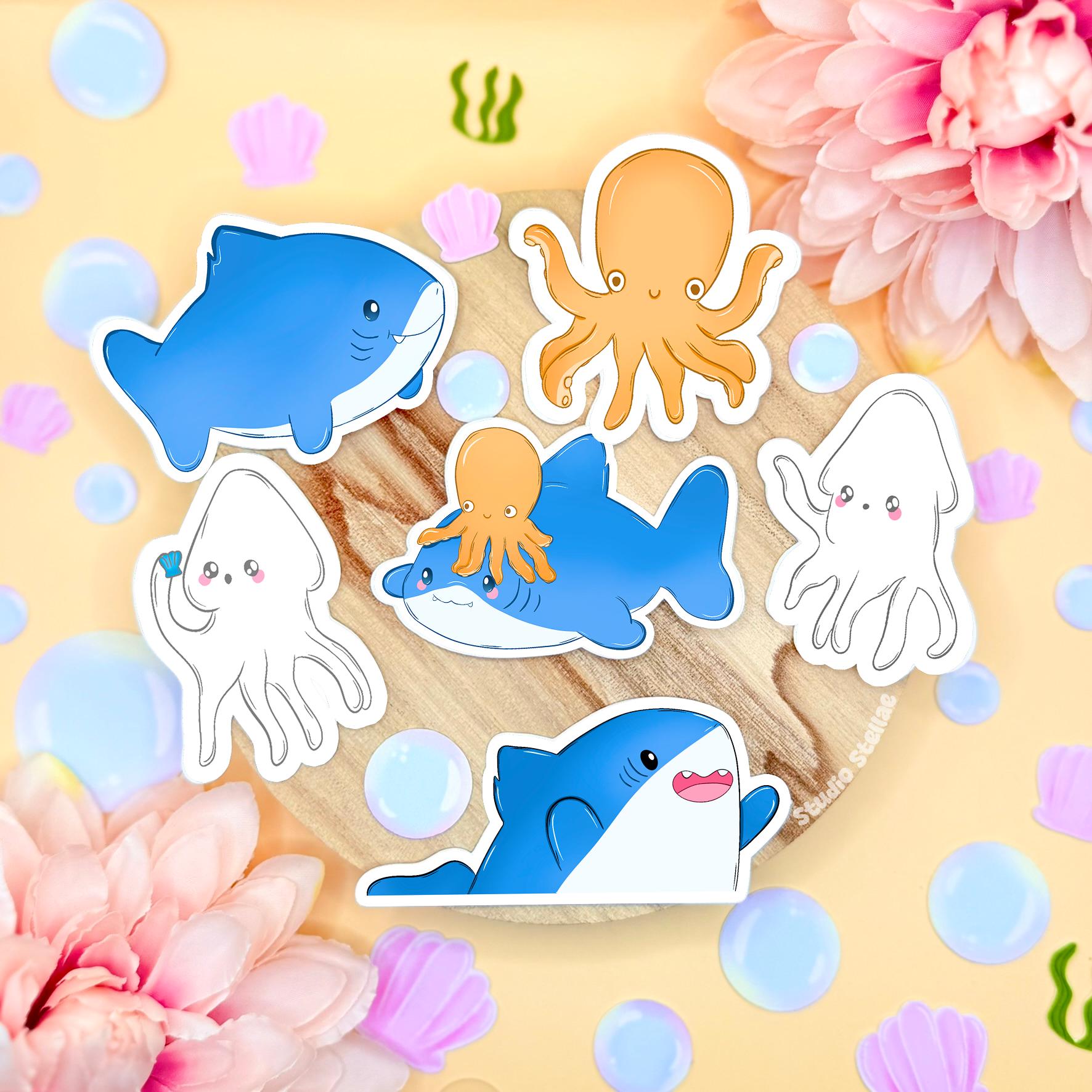 Chibi shark sticker set arranged in a soft pastel flat lay on cork with flowers and bubbles, showing the full kawaii ocean animal sticker pack together. The set includes three blue sharks, two cute squids and one small octopus, all hand drawn in a rounded chibi style. These waterproof matte laminated sea life decals are perfect for decorating laptops, planners, journals and water bottles, and make a sweet gift for ocean lovers.