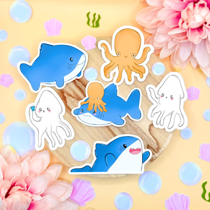 Chibi shark sticker set arranged in a soft pastel flat lay on cork with flowers and bubbles, showing the full kawaii ocean animal sticker pack together. The set includes three blue sharks, two cute squids and one small octopus, all hand drawn in a rounded chibi style. These waterproof matte laminated sea life decals are perfect for decorating laptops, planners, journals and water bottles, and make a sweet gift for ocean lovers.