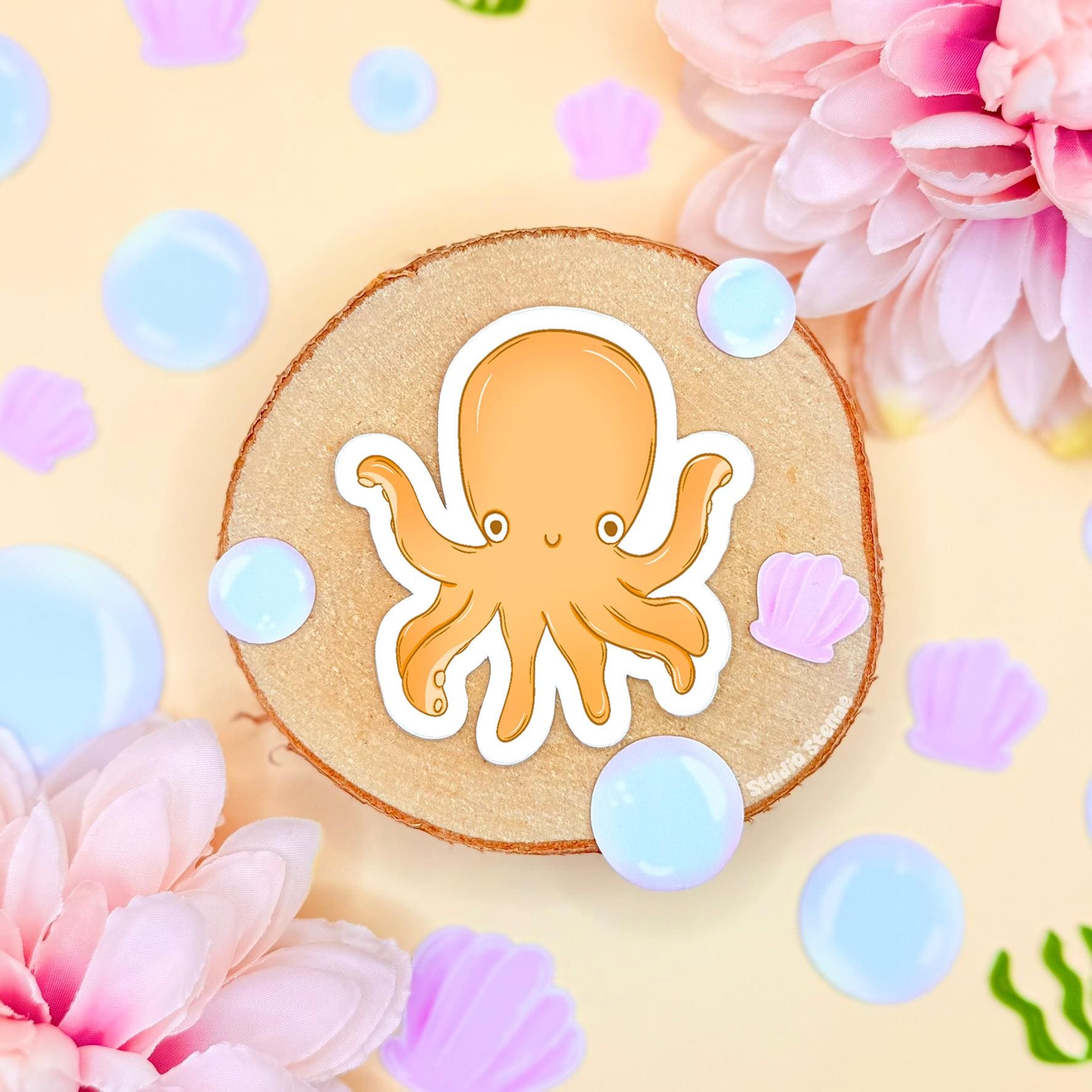 Close-up of the small yellow chibi octopus sticker placed on cork with pastel flowers and bubble accents, showing fine details, smooth matte laminate and clean cut outline. The cheerful ocean creature adds a playful pop of color to planners, journals and water bottles and pairs perfectly with the shark and squid designs in the set, making it a cute and fun sea life decoration.