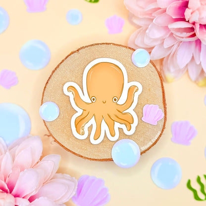 Close-up of the small yellow chibi octopus sticker placed on cork with pastel flowers and bubble accents, showing fine details, smooth matte laminate and clean cut outline. The cheerful ocean creature adds a playful pop of color to planners, journals and water bottles and pairs perfectly with the shark and squid designs in the set, making it a cute and fun sea life decoration.