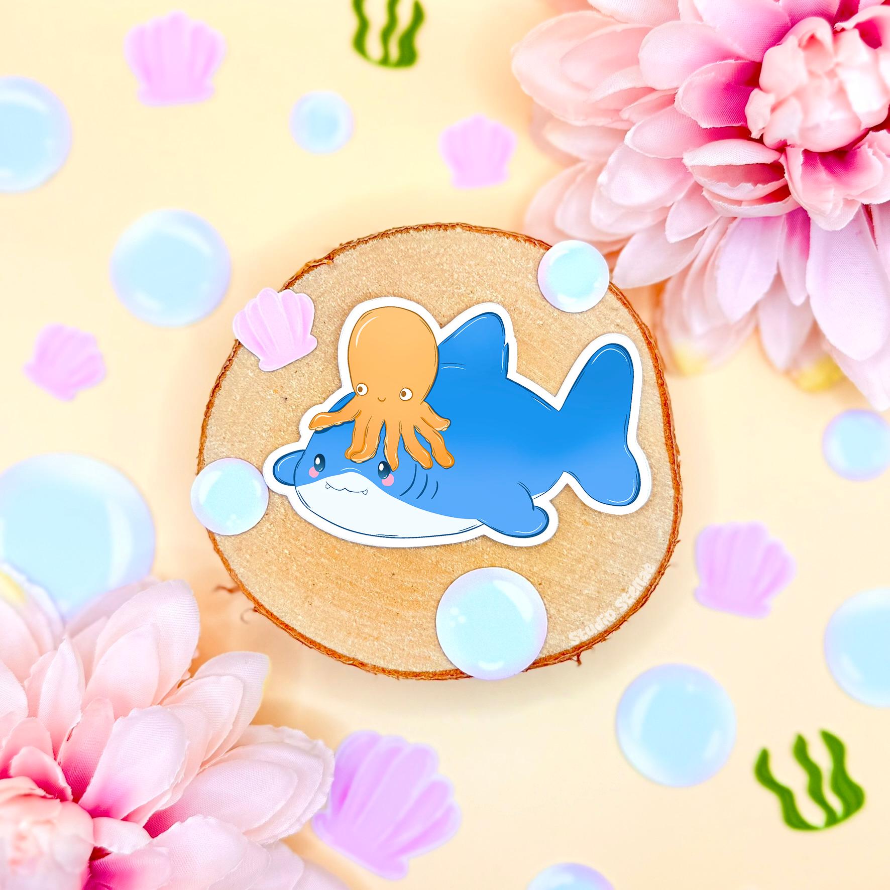 Two ocean animal stickers, including a smiling blue shark and yellow octopus, styled together in a soft pastel flat lay with flowers and cork. Shows how the designs look when paired and highlights their cohesive chibi style and bright colors. Perfect for themed planner spreads, scrapbooking pages, laptops and water bottles, adding playful underwater charm to any stationery setup.