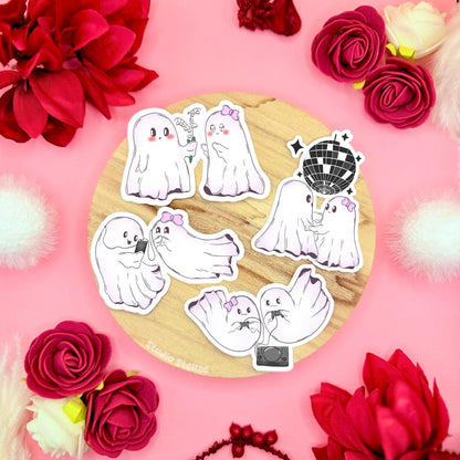 Cute ghost couple sticker set styled in a soft pastel flat lay on cork with red roses and cozy props, showing multiple kawaii ghost characters together. The tiny spooky sweethearts are drawn in a soft chibi style, dancing, cuddling and hanging out with playful expressions. These matte laminated waterproof ghost decals are perfect for decorating laptops, planners, journals and water bottles, and make a sweet Halloween Boo Basket or Valentine gift.