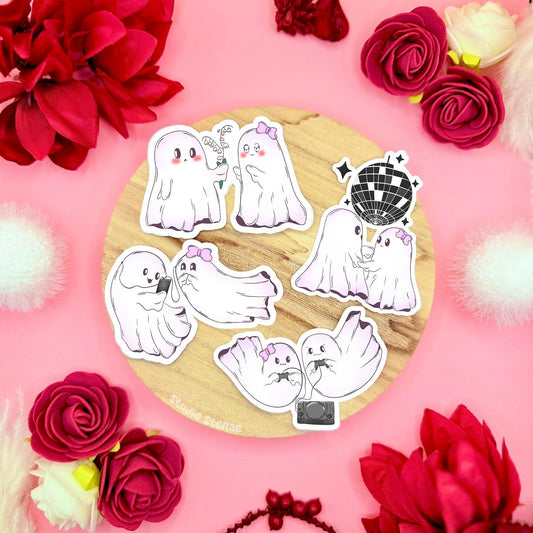 Cute ghost couple sticker set styled in a soft pastel flat lay on cork with red roses and cozy props, showing multiple kawaii ghost characters together. The tiny spooky sweethearts are drawn in a soft chibi style, dancing, cuddling and hanging out with playful expressions. These matte laminated waterproof ghost decals are perfect for decorating laptops, planners, journals and water bottles, and make a sweet Halloween Boo Basket or Valentine gift.