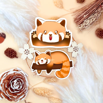 Two cute red panda stickers displayed on a soft beige and floral flat lay, showing both designs from the kawaii chibi sticker pack: one happy, energetic panda and one sleepy, lazy panda curled up. The matte laminated handmade stickers are arranged on warm textured props to highlight their cozy colors and clean die-cut edges. A charming overview of the full red panda sticker set for laptops, planners, journals and water bottles.