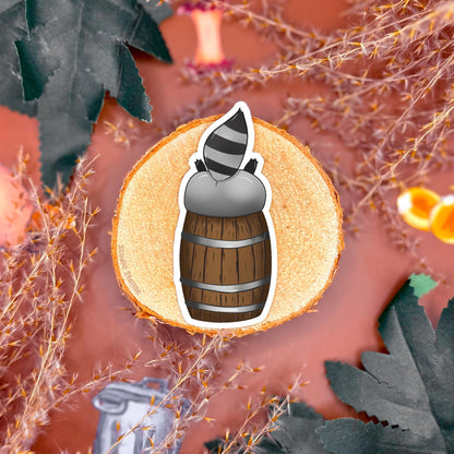 Close up of a rogue raccoon sticker diving headfirst into a wooden barrel with its tail and back legs sticking up, displayed on a warm fantasy styled tabletop with greenery and props. This playful trash panda design captures mischievous DnD energy and chaotic rogue humor, highlighting the matte finish and hand illustrated details. Part of a tabletop RPG raccoon sticker set perfect for decorating dice boxes, laptops, journals, or water bottles and gifting to dungeon masters or fantasy fans.