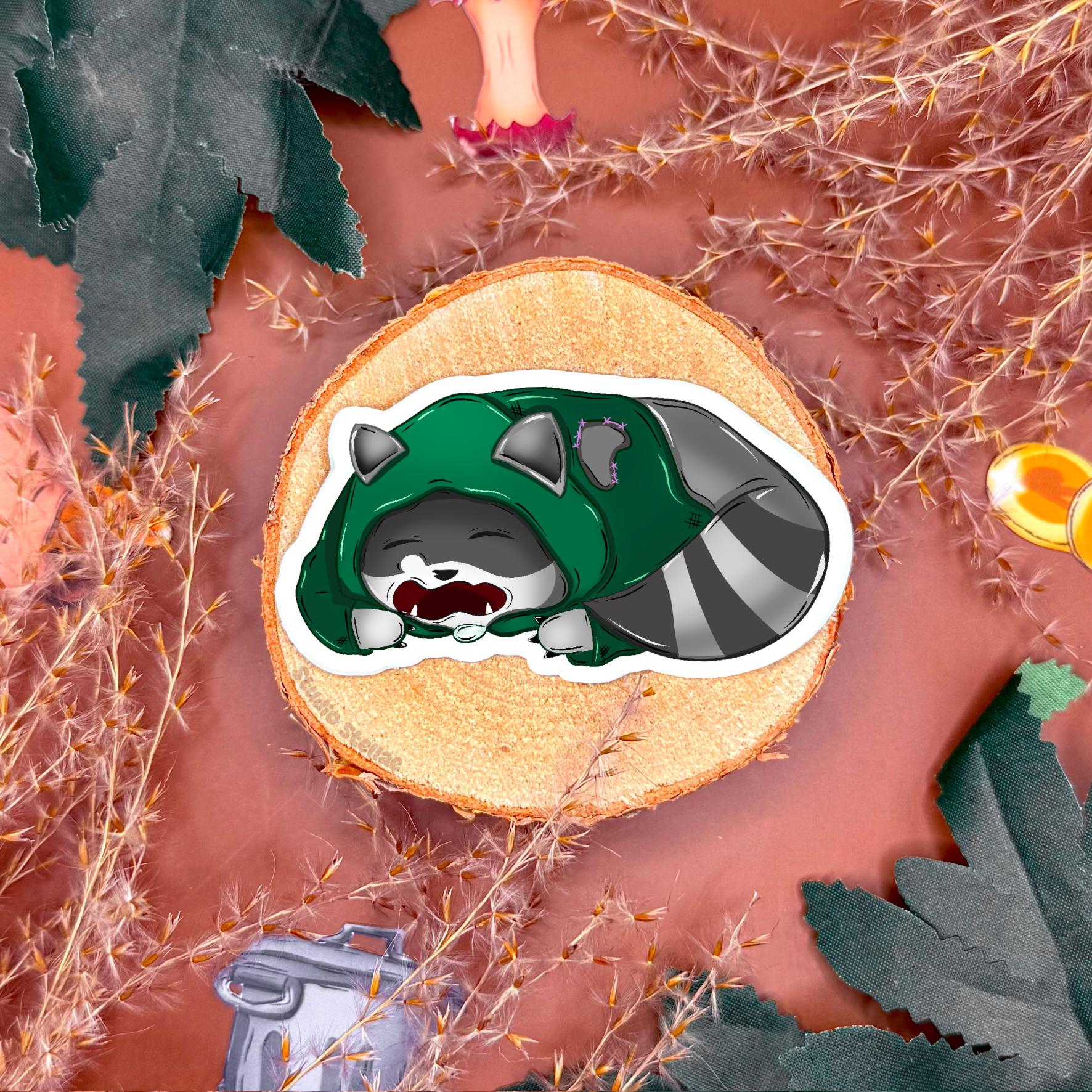 Close up of a sleepy rogue raccoon sticker displayed on a styled tabletop with cozy fantasy props, showing detailed illustration and texture. This image emphasizes the cute chibi design and roleplaying theme of the tabletop RPG sticker set, perfect for gamers who enjoy whimsical fantasy characters.