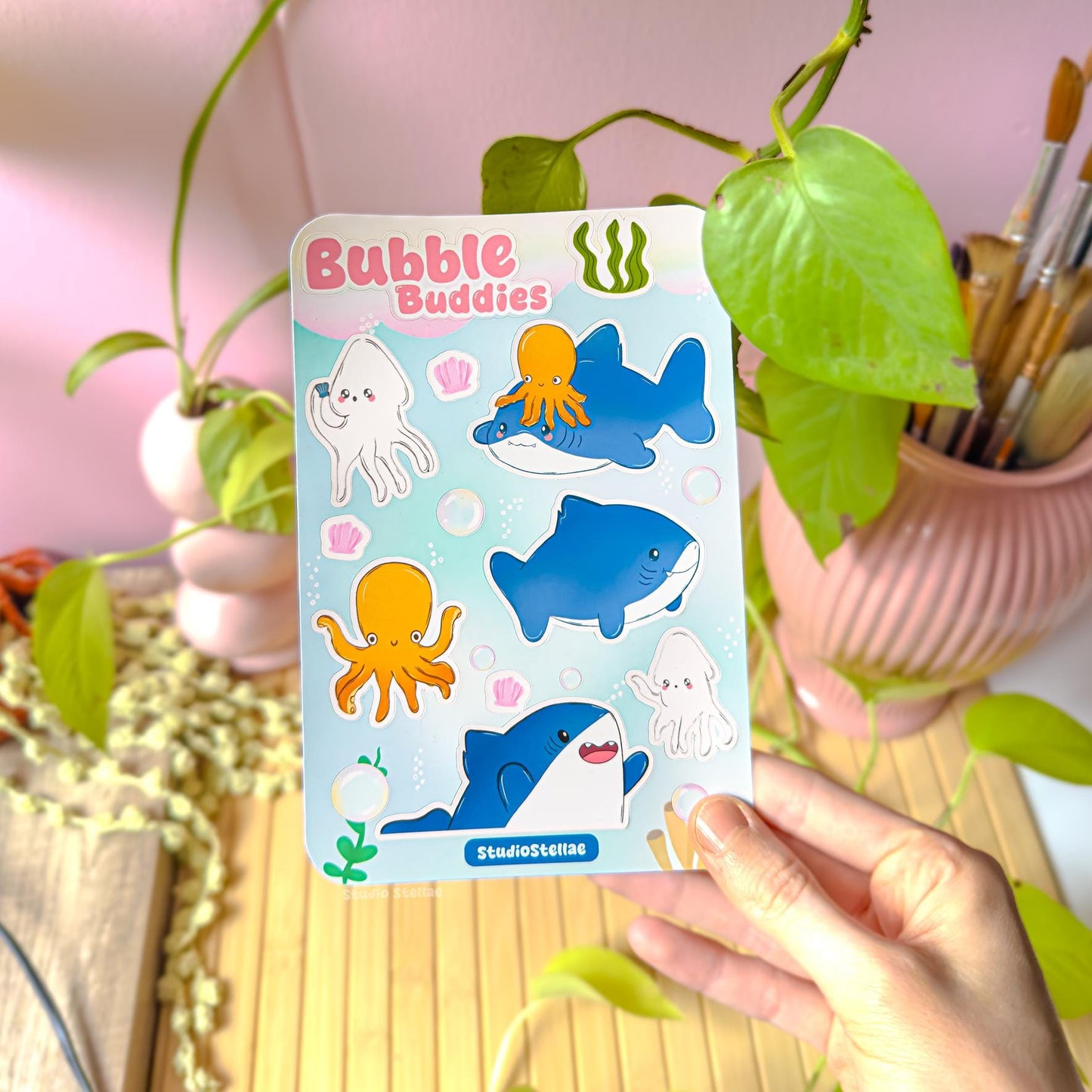 Hand holding the chibi shark sticker sheet near a leafy plant in soft natural light, demonstrating scale, colors, and finish. The cute sharks, squid, octopus, and bubble elements are clearly visible, helping buyers imagine using the sticker sheet to decorate journals, planners, water bottles, or laptops while showing how vibrant the artwork looks in real life.