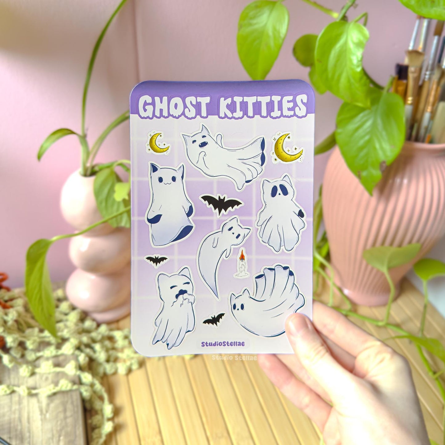 Hand holding the Ghost Kitties sticker sheet in a cozy workspace with green plant leaves and soft natural light. The white ghost cats, crescent moons, and bats stand out against a pastel purple design. This lifestyle photo shows real-life scale and the cute spooky Halloween aesthetic, perfect for witchy vibes, dark academia decor, and cat lover gifts.