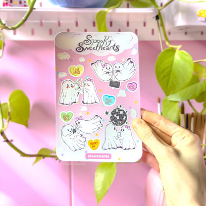 Hand holding the Spooky Sweethearts sticker sheet against a soft pink background with green plant leaves. The full sheet shows ghost couples in sweet moments surrounded by candy hearts and clouds. This lifestyle photo shows real-life scale and how the stickers look in natural light, perfect for journaling, scrapbooking, or decorating laptops and planners.
