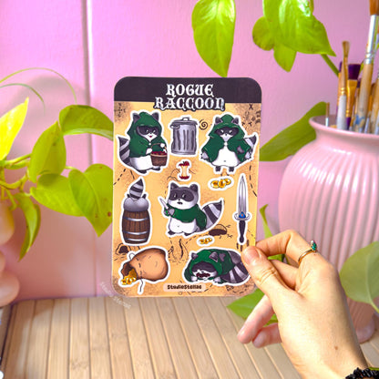 Hand holding the Rogue Raccoon sticker sheet in a cozy workspace with green plant leaves and a pink vase in soft natural light. The full sheet displays raccoon rogue characters, coins, treasure items, and fantasy accessories. This lifestyle photo shows real-life scale and how the stickers look in hand, perfect for DnD players, TTRPG fans, journaling, planners, and laptop decoration.