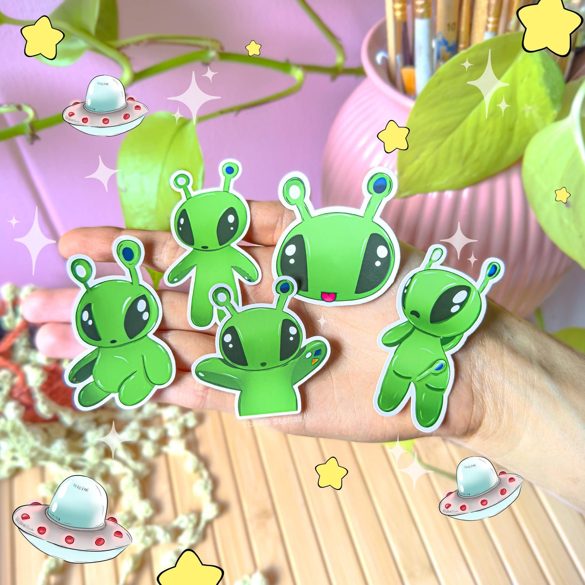 All five Aftonsparv alien stickers displayed together on a wooden desk beside trailing green plants, photographed in natural light to show the complete 5-piece set. The bright green kawaii alien characters create a playful, cohesive collection perfect for decorating planners, laptops, journals and water bottles. Demonstrates how the full sticker pack looks grouped together in a cozy workspace.