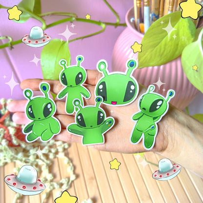 All five Aftonsparv alien stickers displayed together on a wooden desk beside trailing green plants, photographed in natural light to show the complete 5-piece set. The bright green kawaii alien characters create a playful, cohesive collection perfect for decorating planners, laptops, journals and water bottles. Demonstrates how the full sticker pack looks grouped together in a cozy workspace.