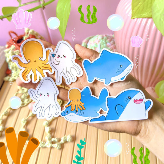All ocean animal stickers displayed together on a wooden desk beside trailing green plants, photographed in natural daylight to show the complete 6-piece chibi shark sticker set. The mix of smiling sharks, squids and octopus creates a playful underwater themed collection. Perfect for decorating planners, scrapbooks, laptops and water bottles with cute sea life decals that brighten any cozy workspace.