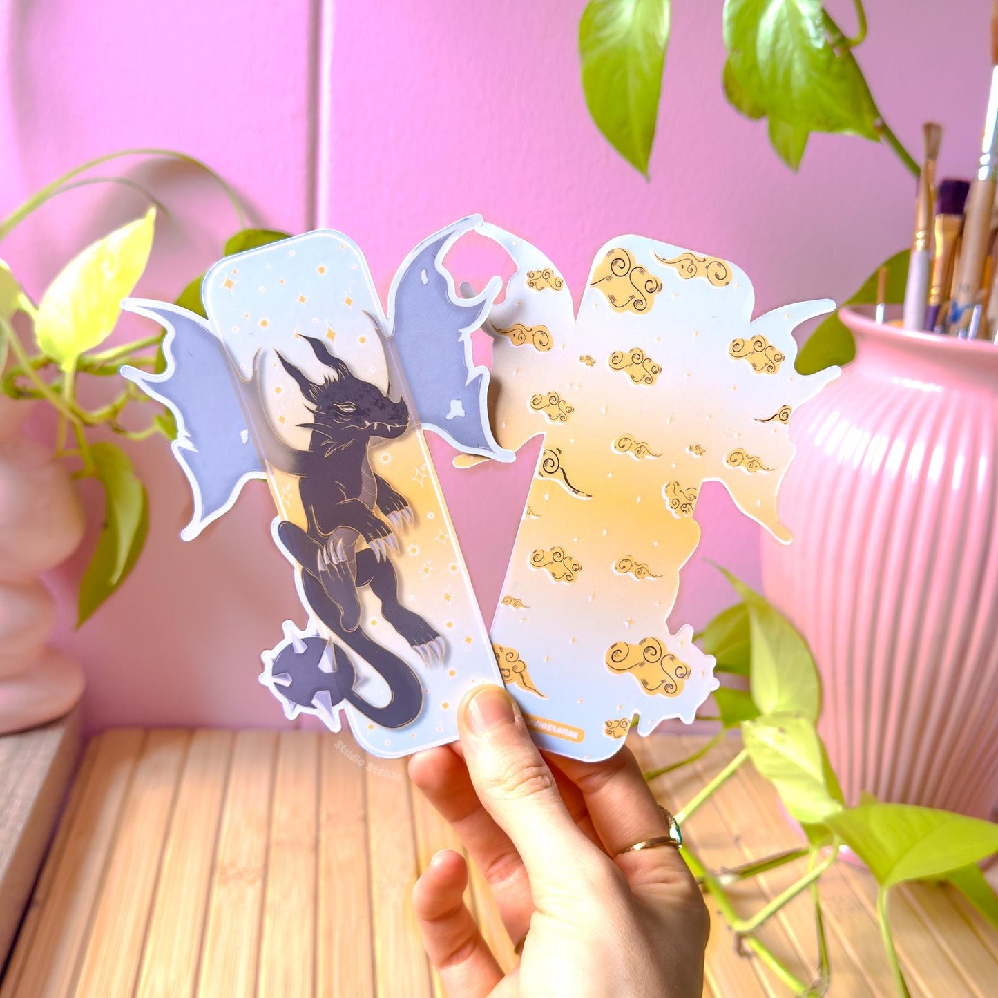 Hand holding the pastel Fourth Wing inspired dragon bookmark in a cozy studio setup with pink wall, green plant leaves, and a pink vase in the background. The fantasy page marker features a shadow dragon silhouette, golden clouds, and starry details in soft pastel colors. This romantasy bookmark is shown in natural light to highlight the smooth matte lamination, sturdy feel, and unique die-cut dragon wing shape for BookTok readers and book lovers.